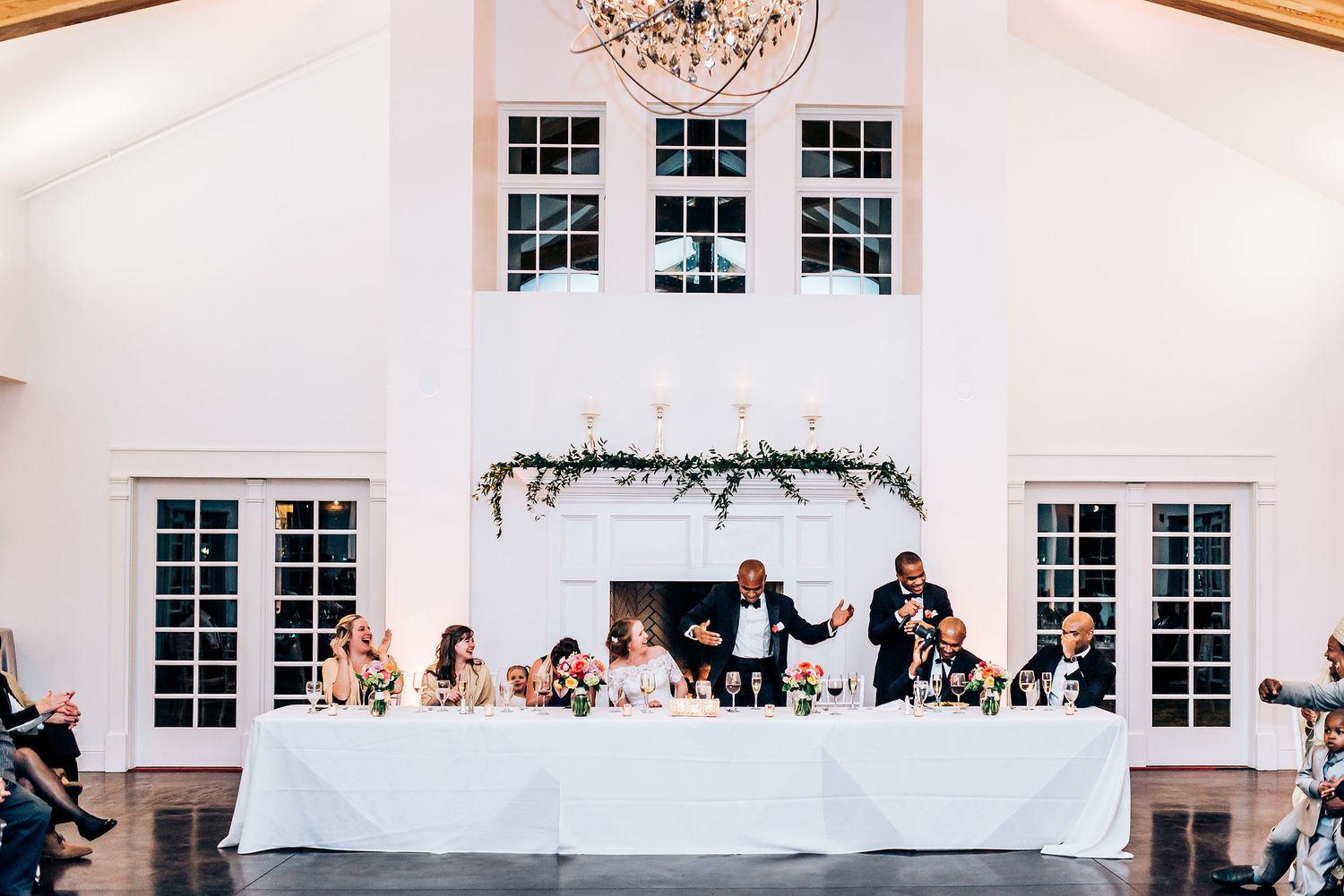 Wedding party seated at long head table under elegant white decor and windows.