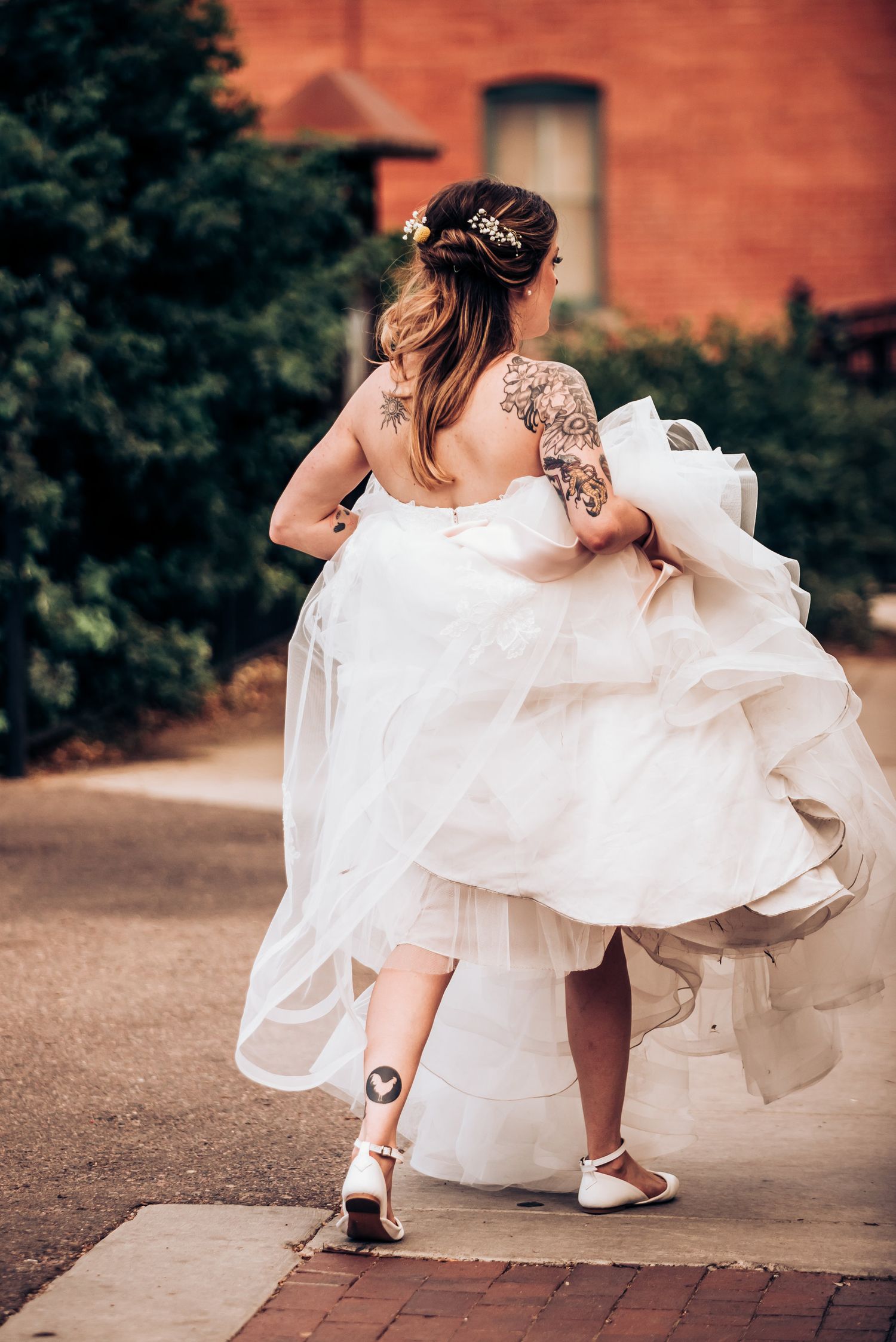 Back view of bride in flowing white dress showing tattoo details while walking on brick path.