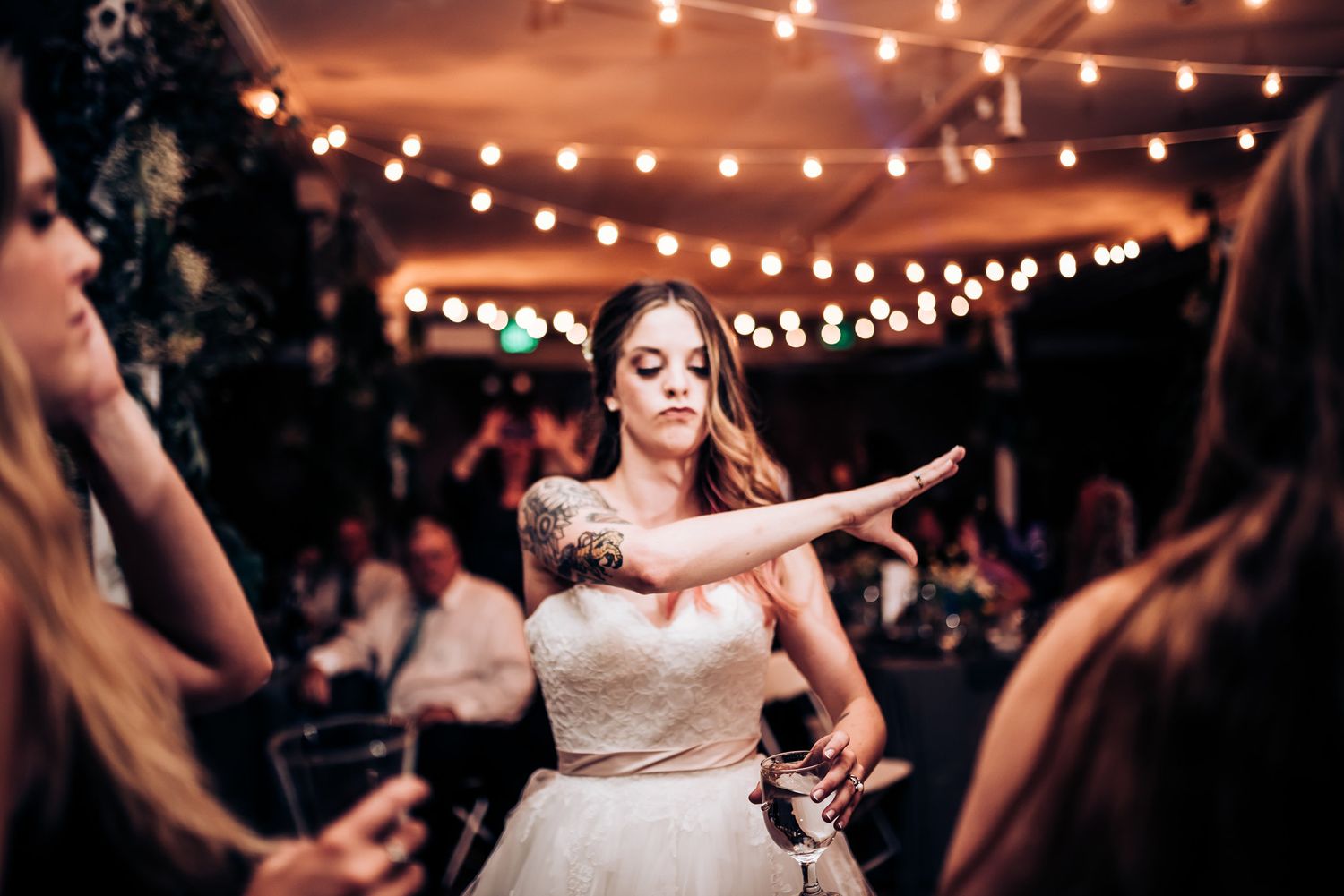 Bride dances energetically among guests under string lights at indoor reception.