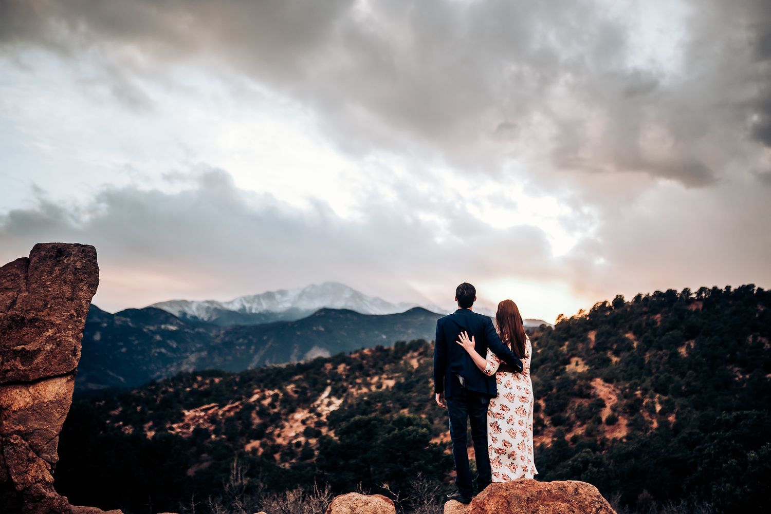 A silhouetted couple stands on a rocky mountain overlook at dusk.