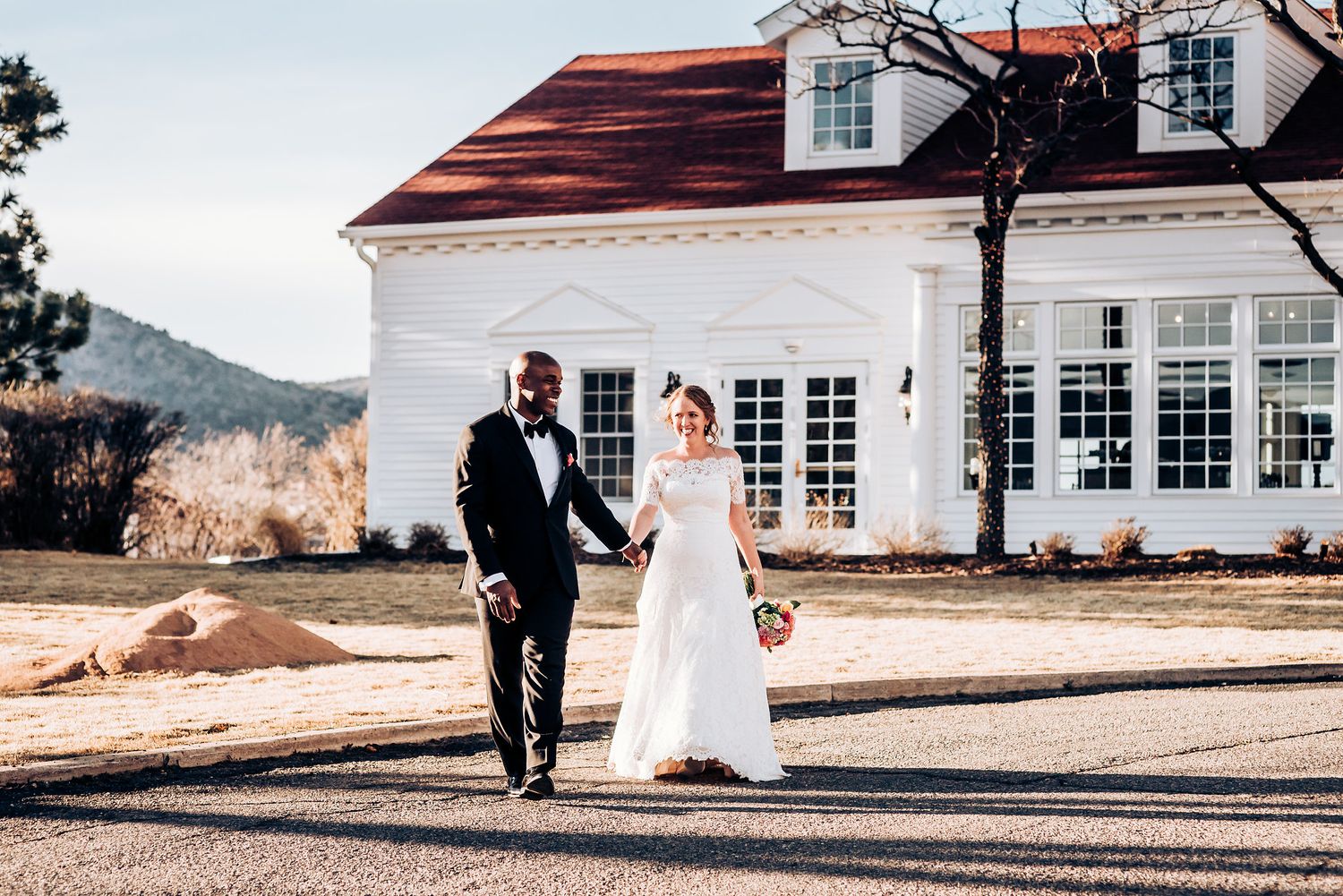 Wedding couple poses in front of white colonial-style house with red roof.