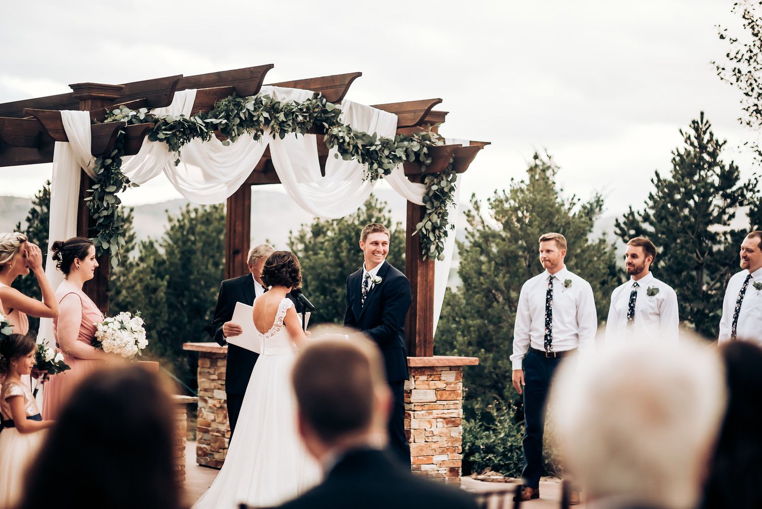A wedding ceremony unfolds beneath a decorated pergola with white draping and greenery.