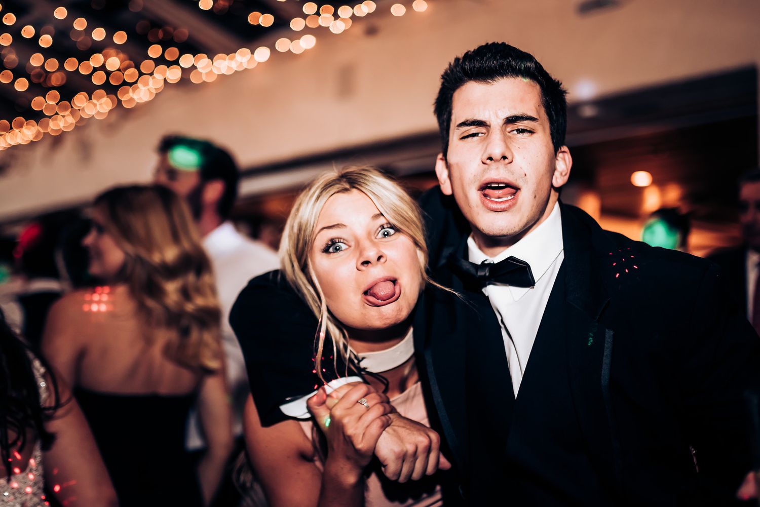 A couple makes silly expressions while dancing at a formal event with string lights overhead.