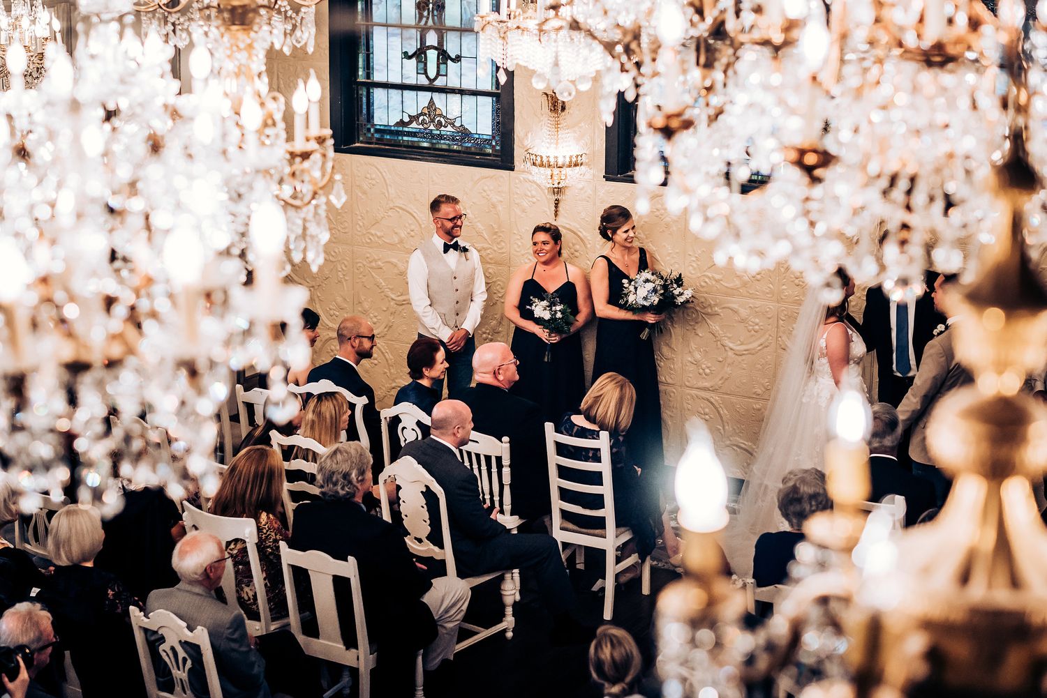 Wedding ceremony captured through hanging crystal chandeliers with guests seated below.