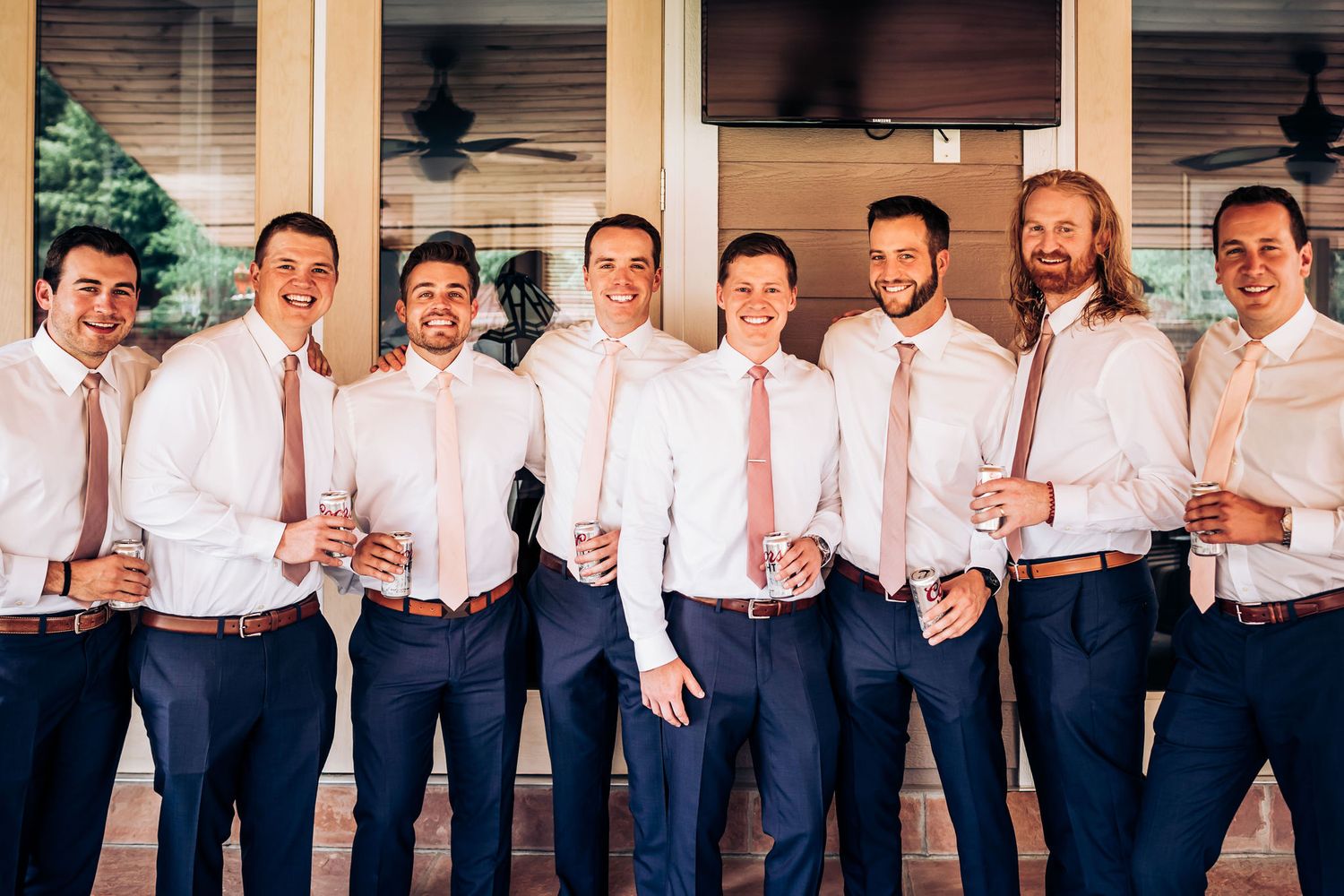 Groomsmen in matching white shirts and pink ties pose on a porch during a wedding celebration.