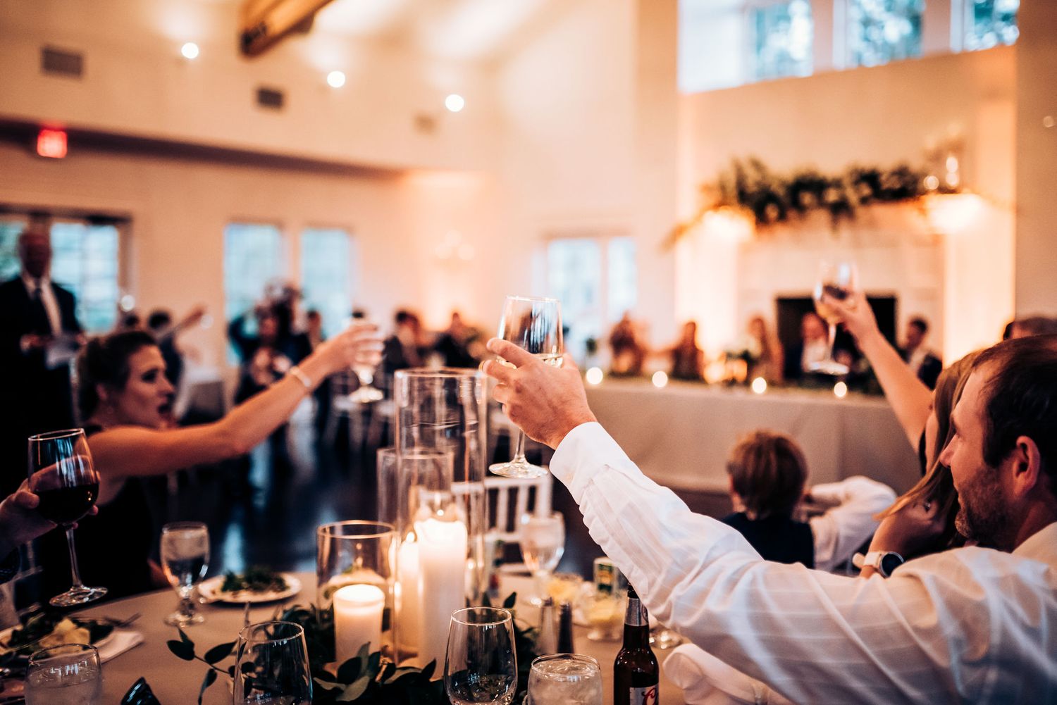 Wedding guests raise glasses for a toast during an elegant reception with candlelit tables.