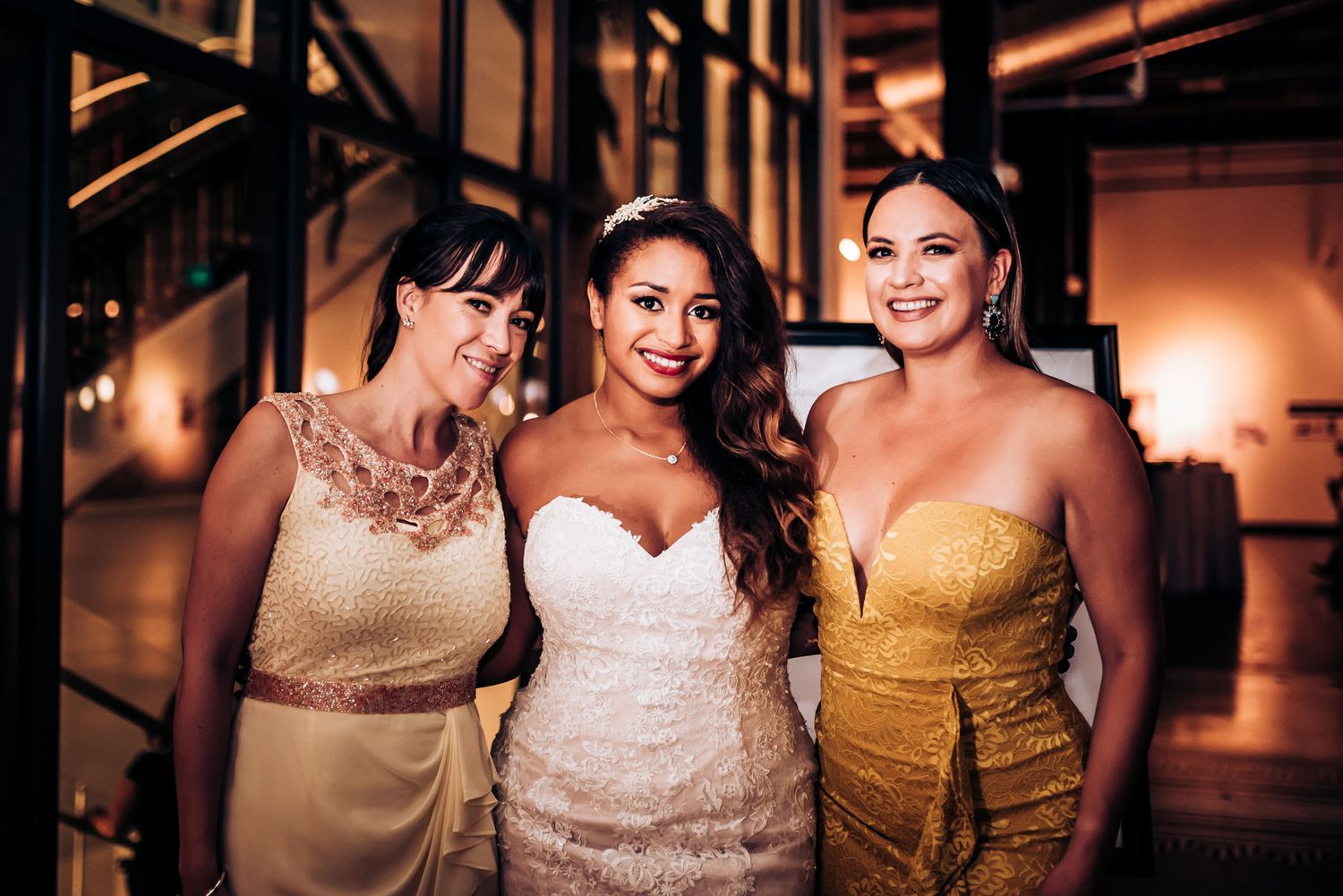 Three women in elegant dresses pose together at a formal wedding celebration.