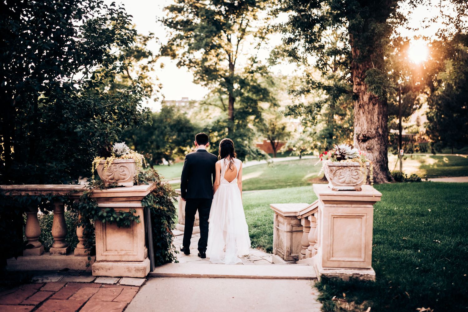 A couple stands together at sunset on elegant stone steps surrounded by lush gardens.