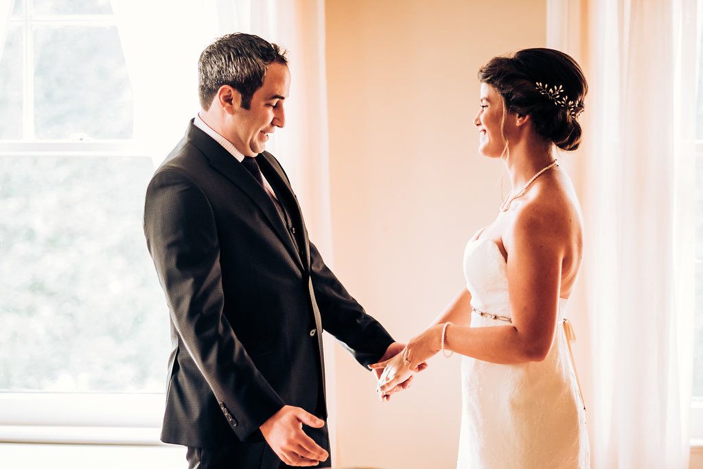 A couple holds hands and smiles during their first look by a bright window.