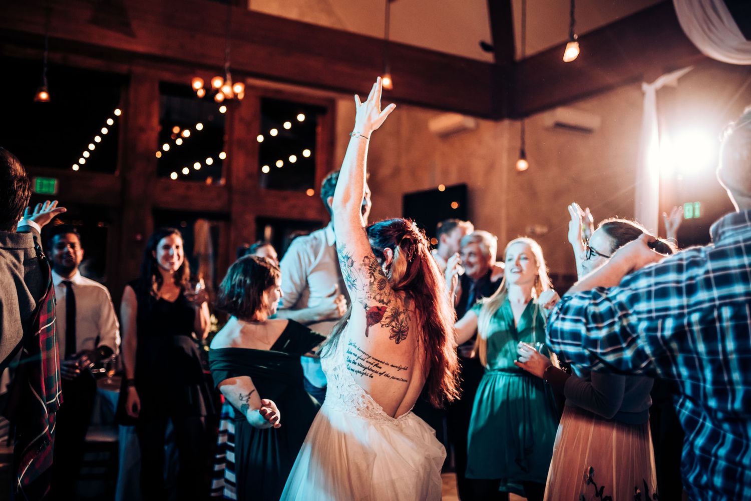 Wedding guests celebrate on the dance floor under warm string lights in a rustic venue.