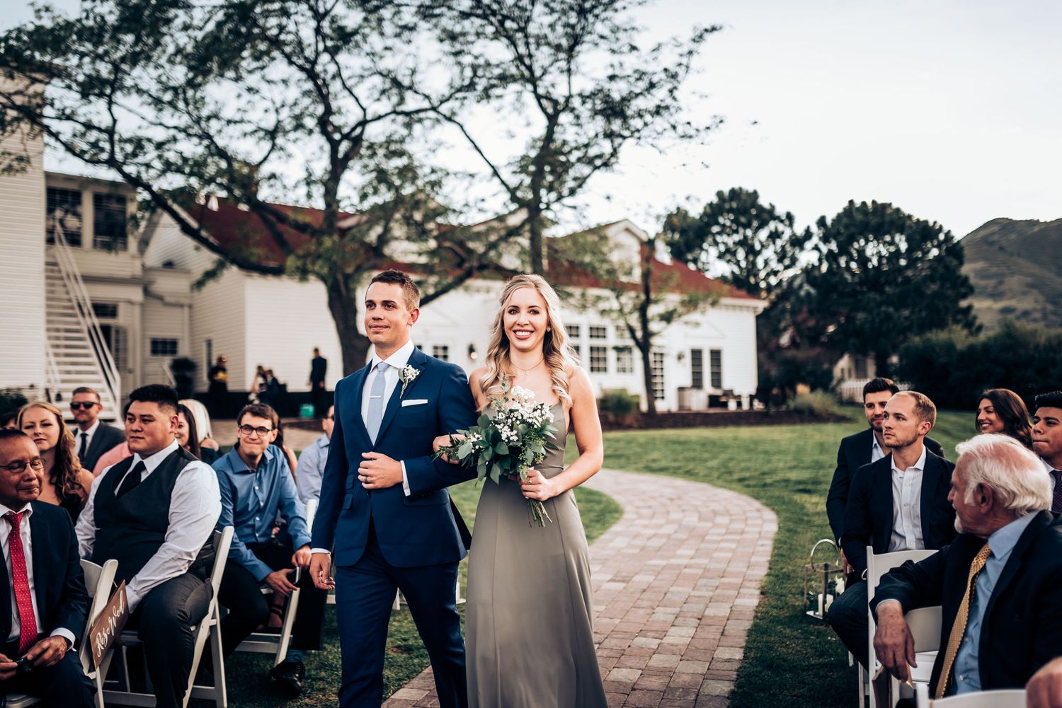 Couple proceeding down brick pathway during outdoor wedding ceremony near white colonial building.