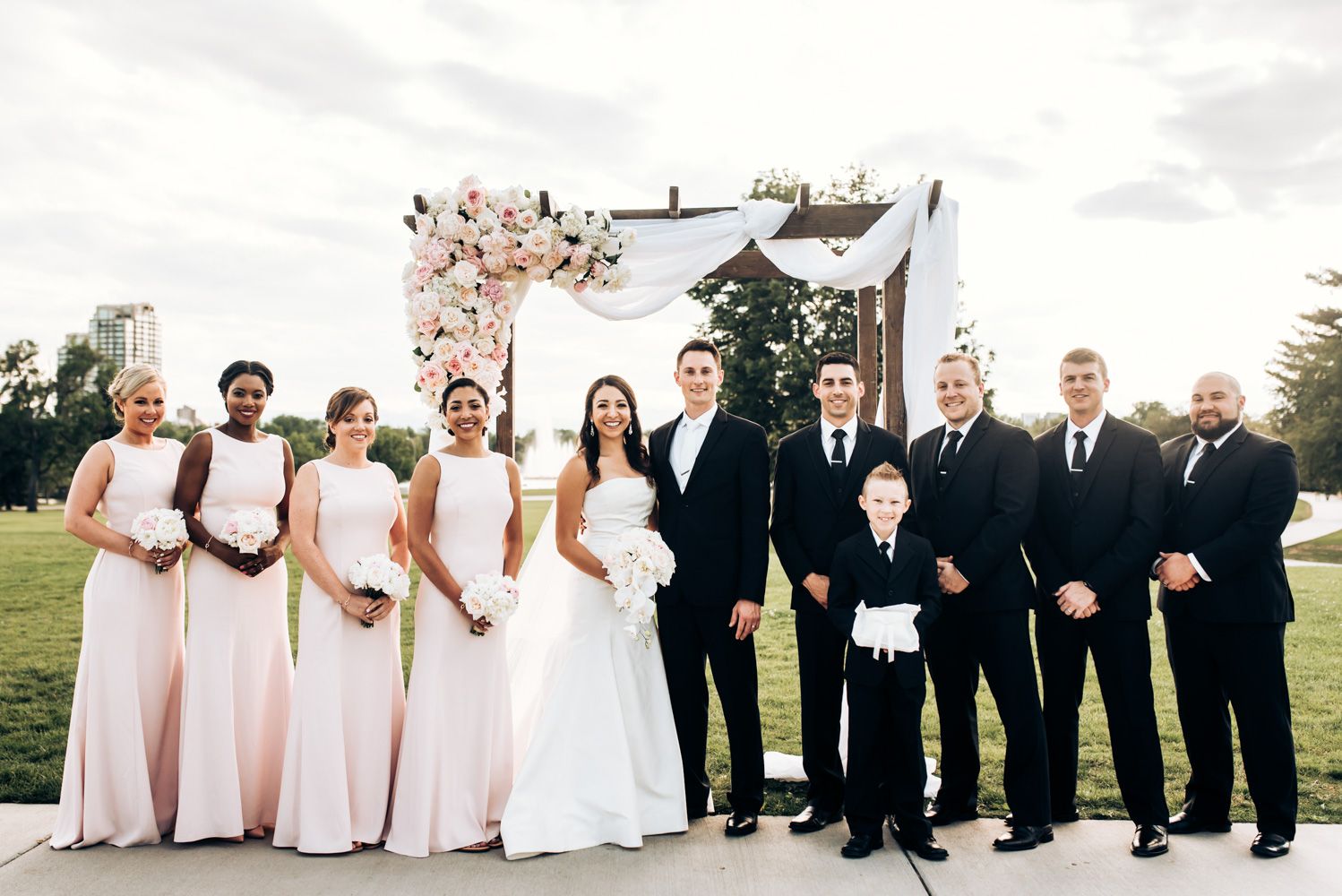 Wedding party group photo with people in pink dresses and black suits under a floral arch.