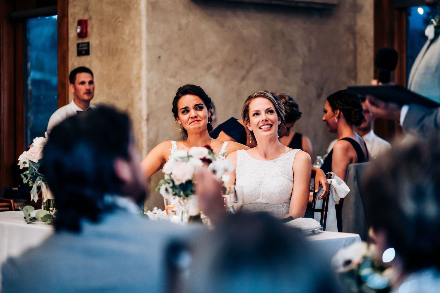 Wedding reception guests gathered around decorated tables in elegant venue with natural lighting.