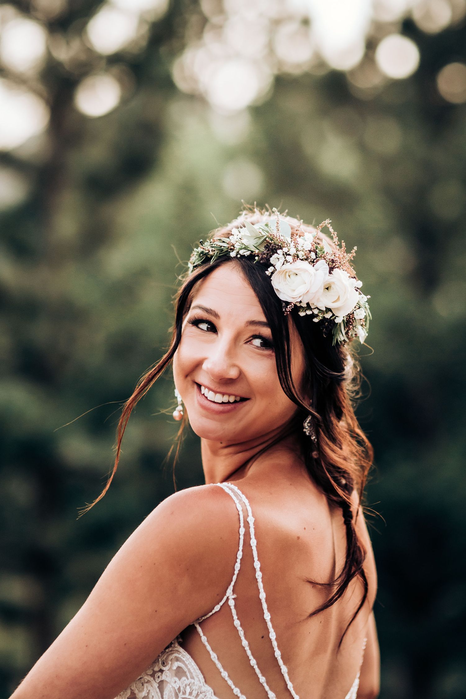 Bride in backless wedding dress with floral crown smiles over shoulder in outdoor portrait.
