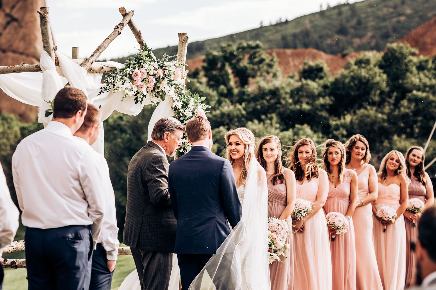 Wedding ceremony under rustic wooden arch with bridal party in pink dresses and mountain landscape backdrop.