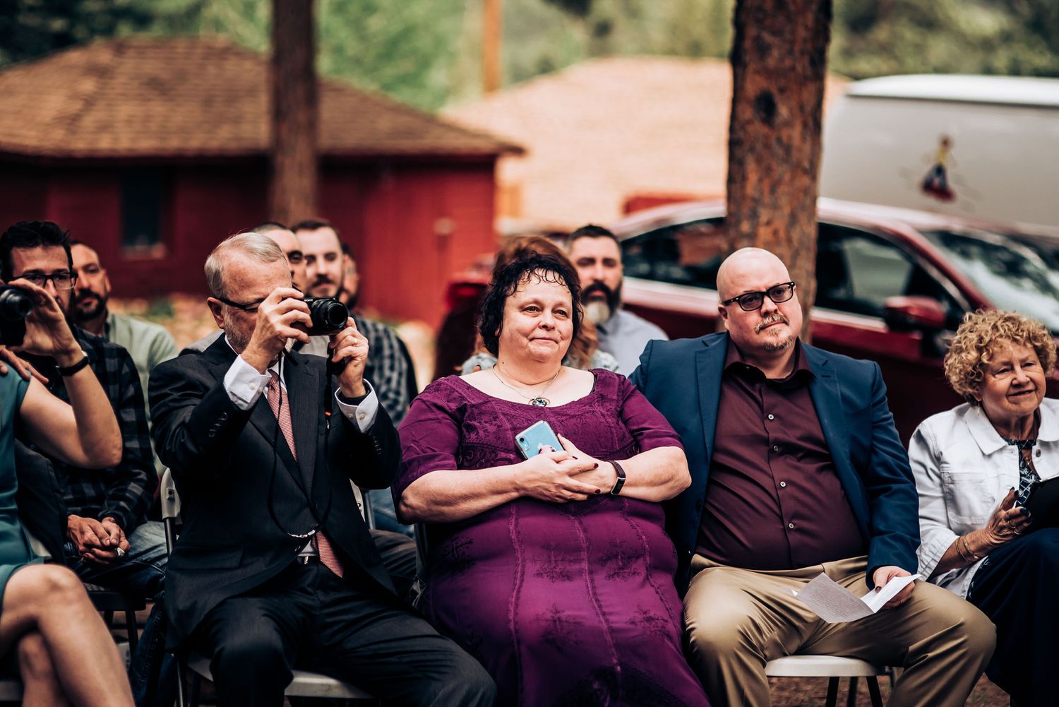 Wedding guests seated outdoors watch an emotional ceremony among pine trees.