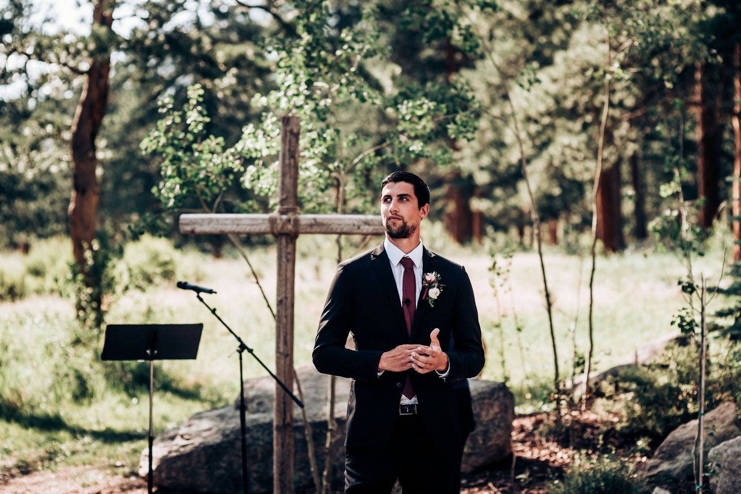 A person in a black suit stands at a wooden altar in an outdoor forest ceremony setting.