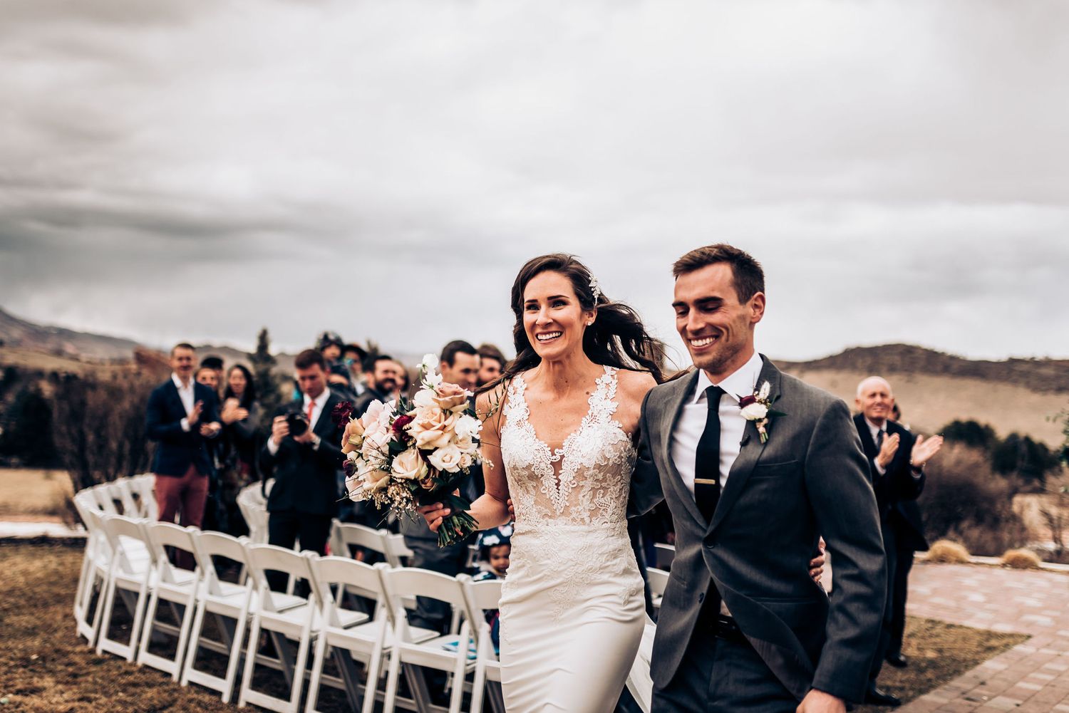 Joyful newlyweds walk down aisle with white chairs and mountain scenery behind them.