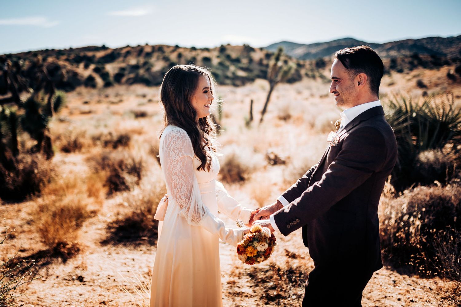 Couple in wedding attire sharing a moment in a desert setting with mountains in background.
