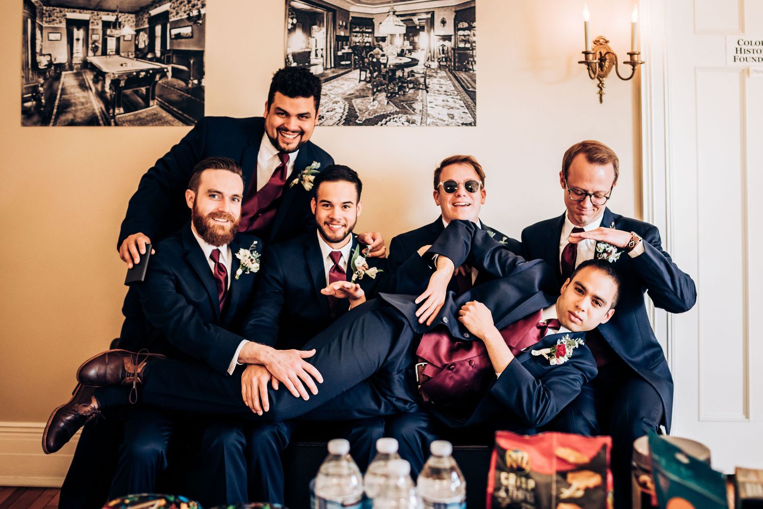 Groomsmen in navy suits lounging casually on a couch while sharing drinks and laughing together.