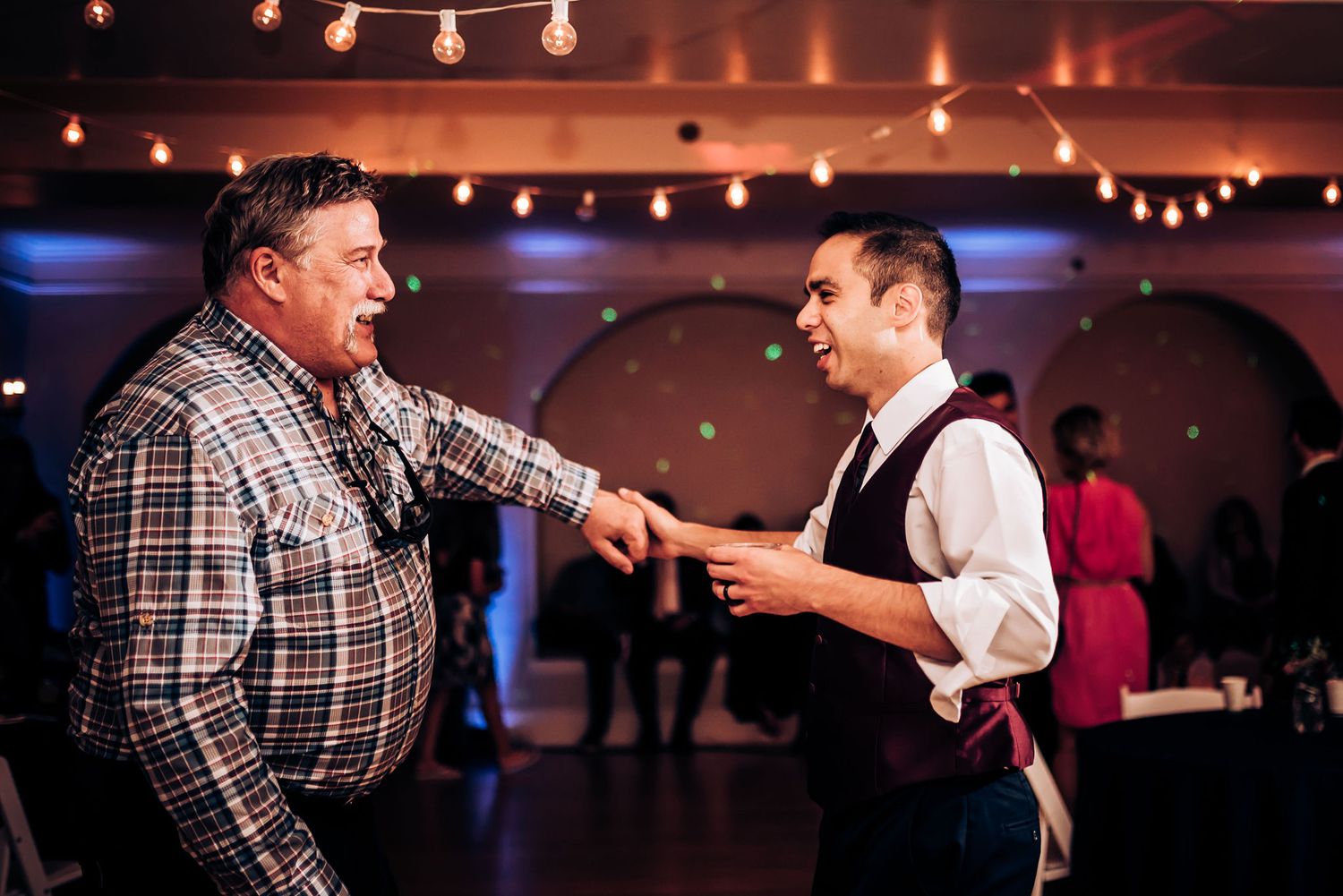 Two people share a playful dance moment at an indoor reception venue with ambient string lights.