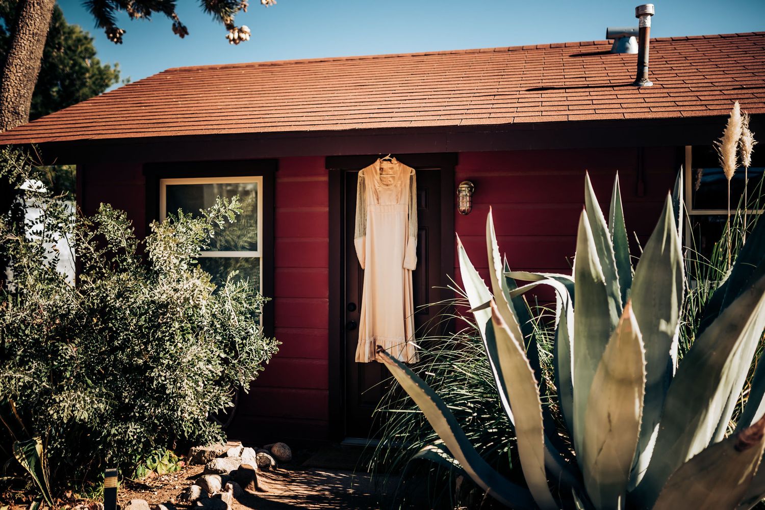 A wedding dress hanging outside a red rustic house with desert plants.