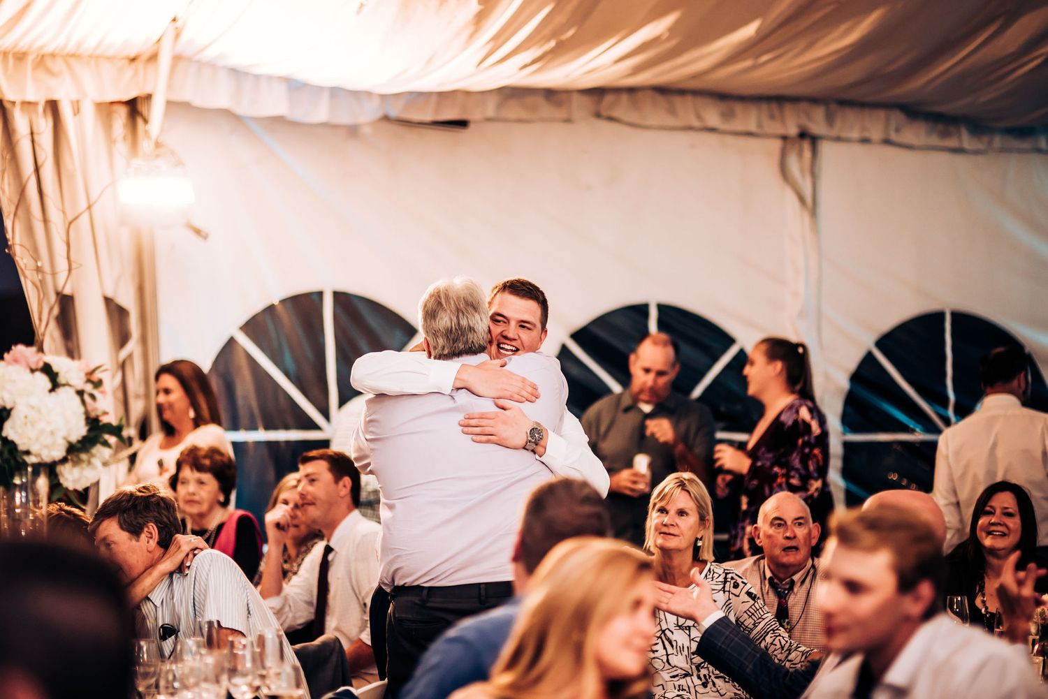 Wedding reception guests celebrate joyfully at tables under a white tent with window panels.