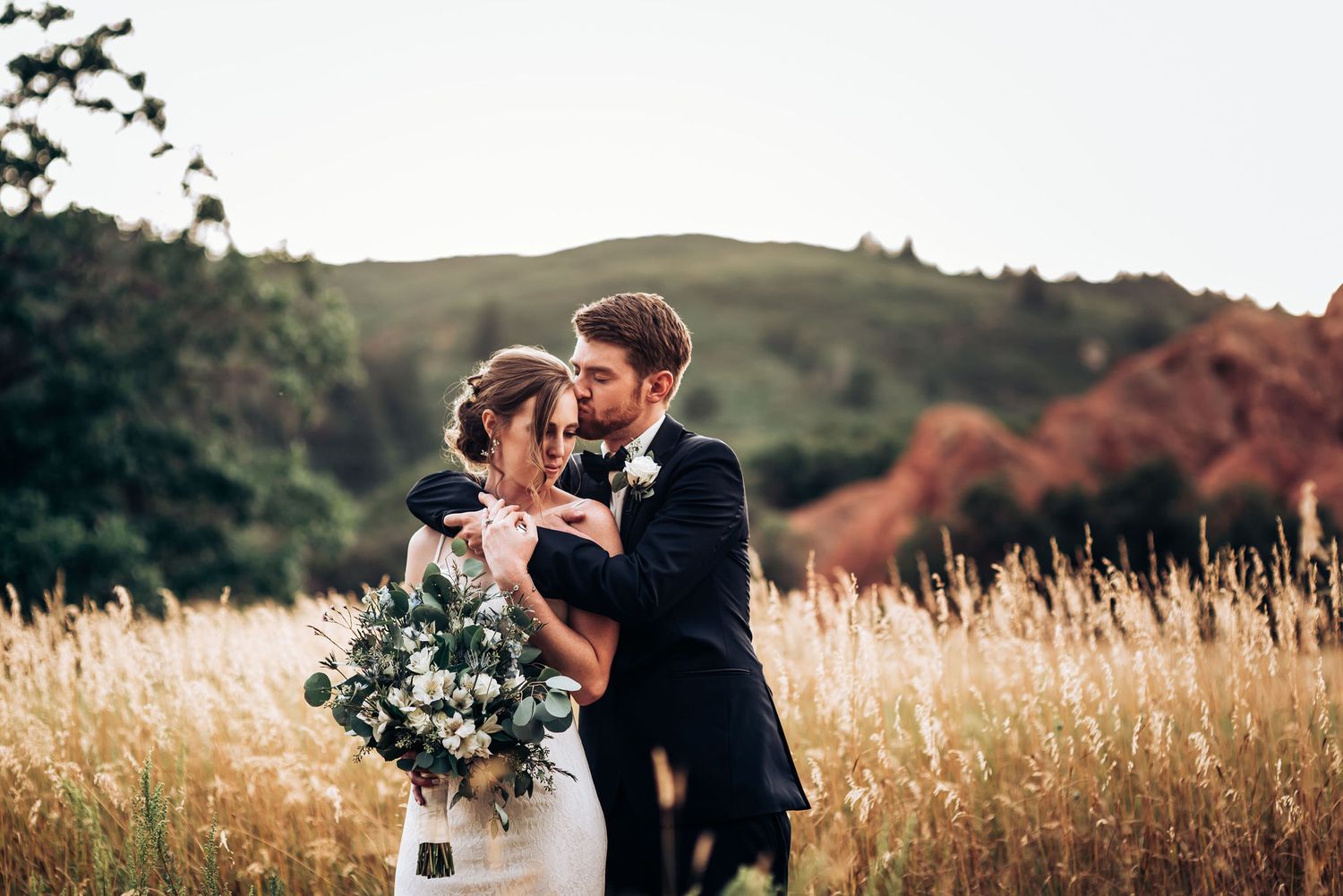 Newlyweds embrace in sunset-lit meadow with romantic mountain backdrop.