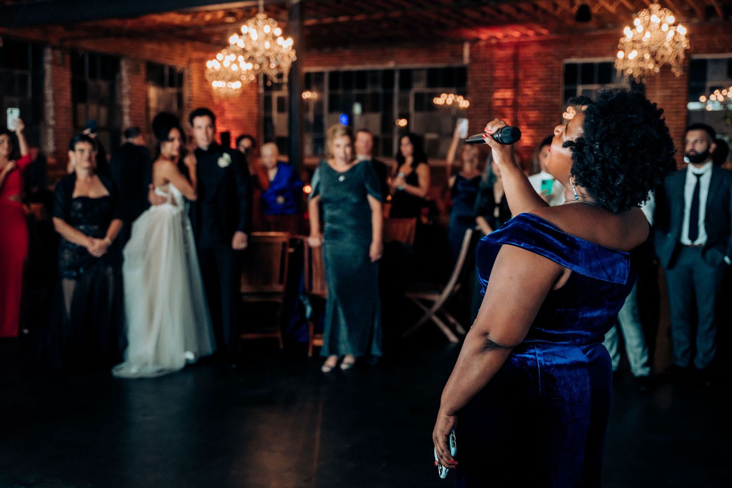 Wedding singer in blue dress performs for guests in an elegant brick-walled venue.