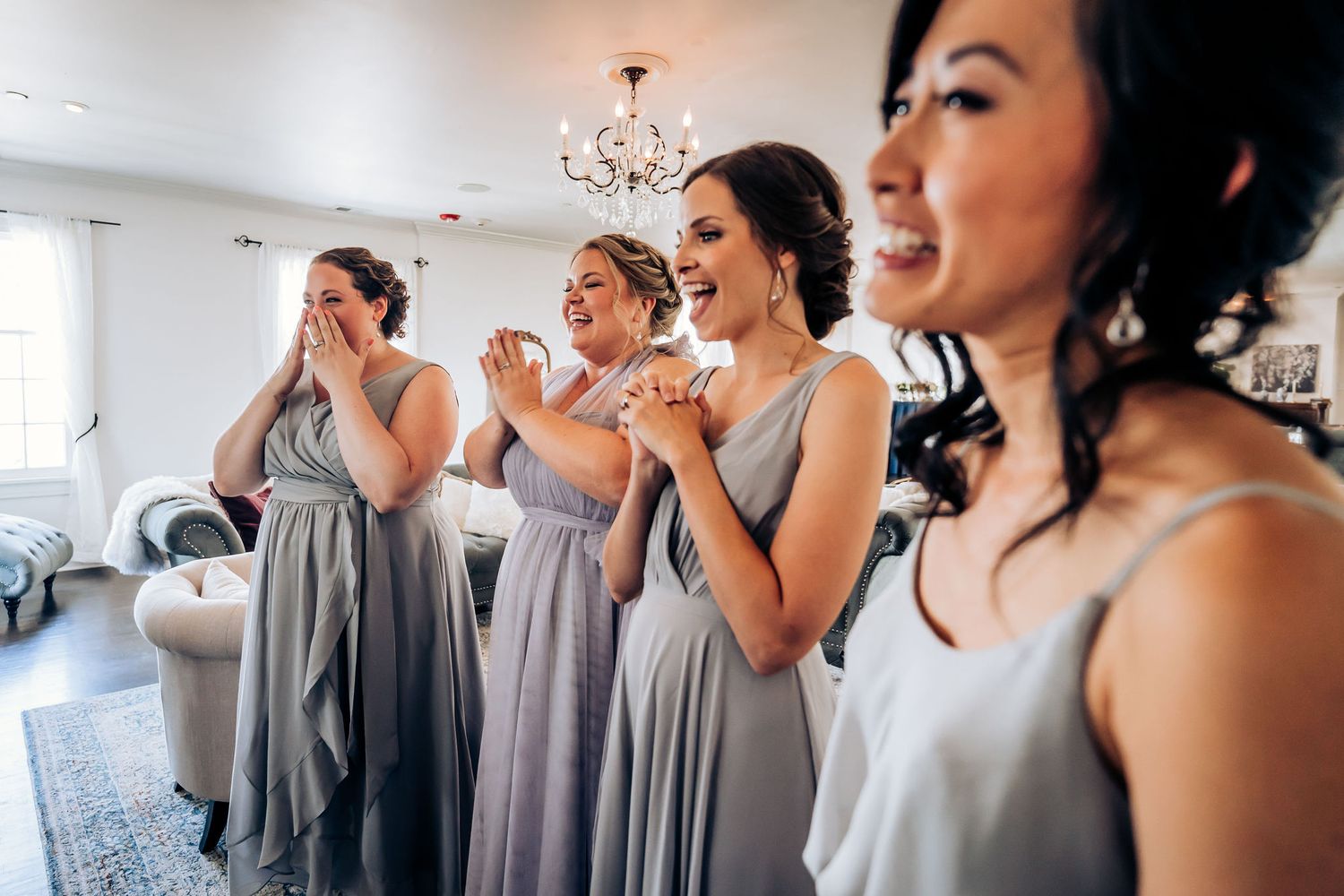 Bridesmaids in matching grey dresses react with joy and emotion during pre-wedding preparations.