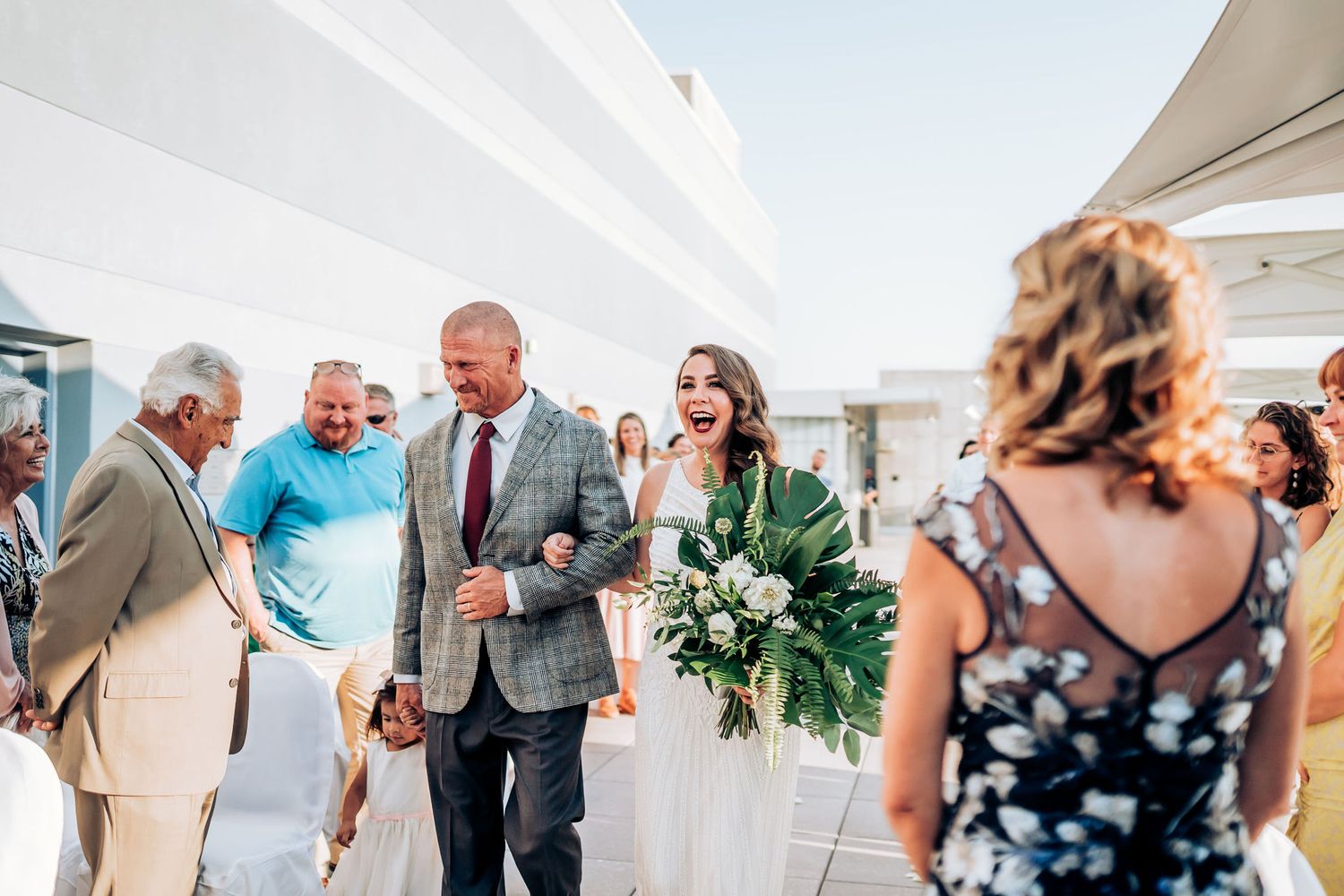 Wedding guests gather on a sunny outdoor terrace with white walls sharing joyful moments during a celebration.