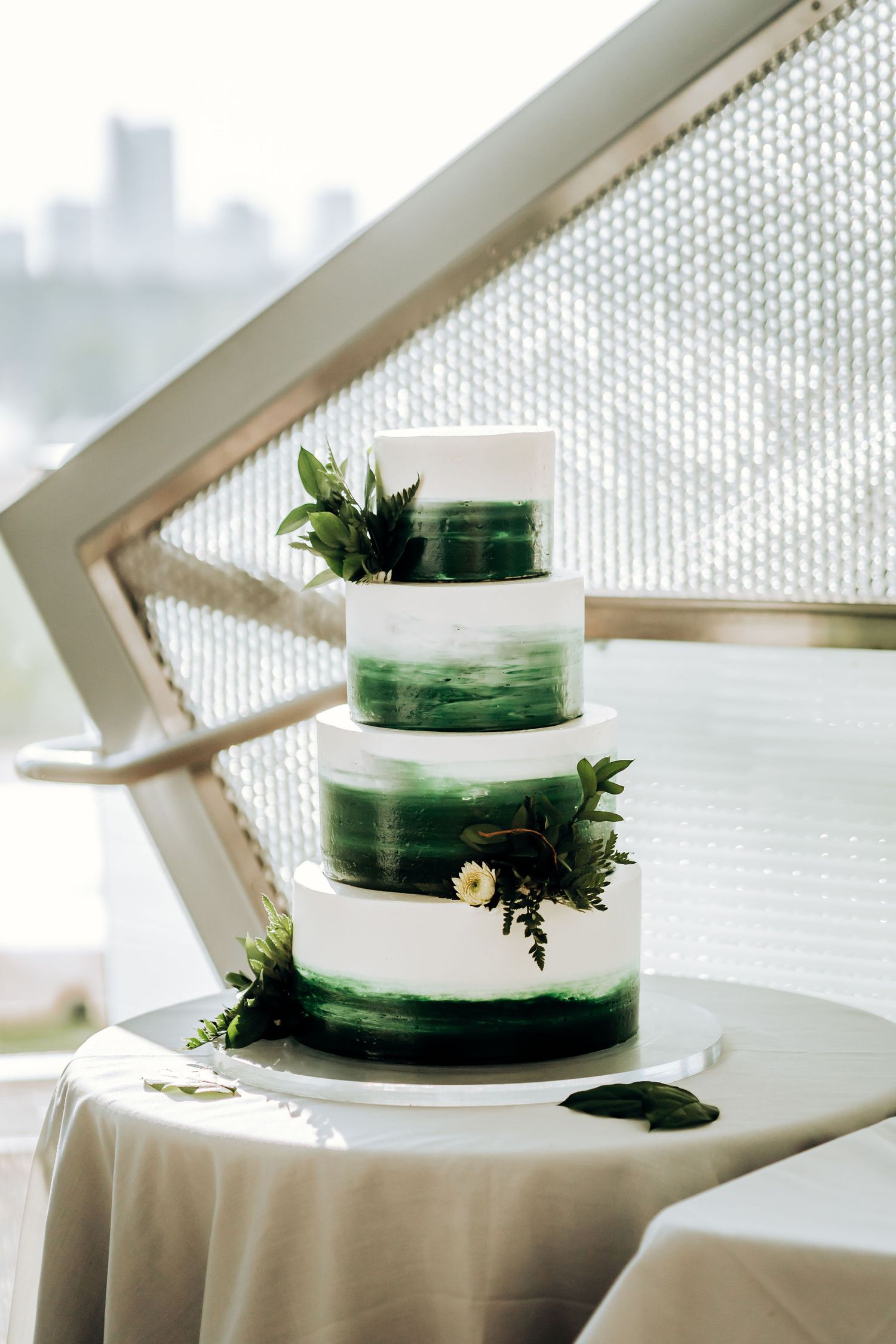 A modern four-tier wedding cake with white and emerald green ombre design decorated with fresh greenery and flowers.