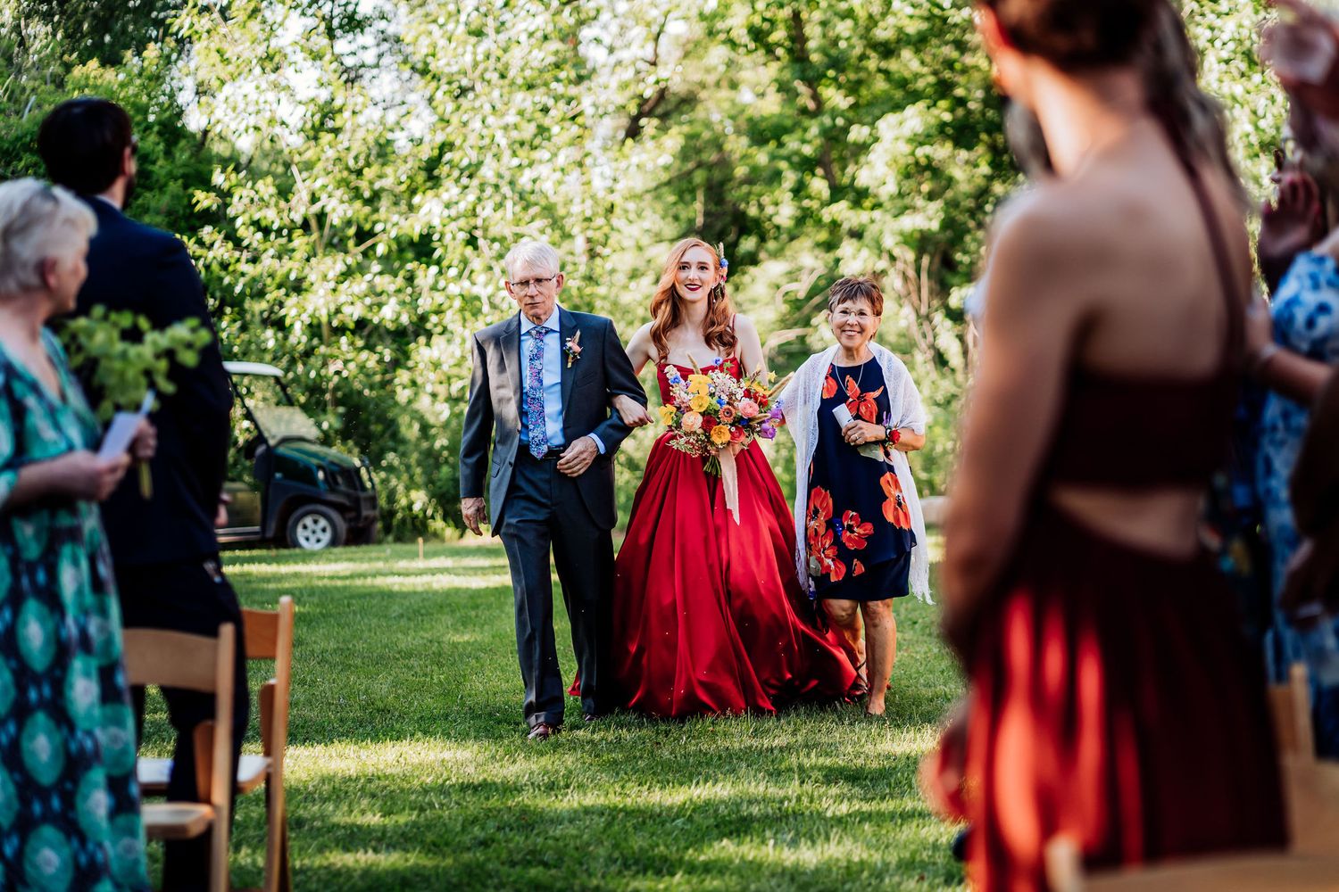 Wedding party walks down an outdoor aisle surrounded by guests and greenery.