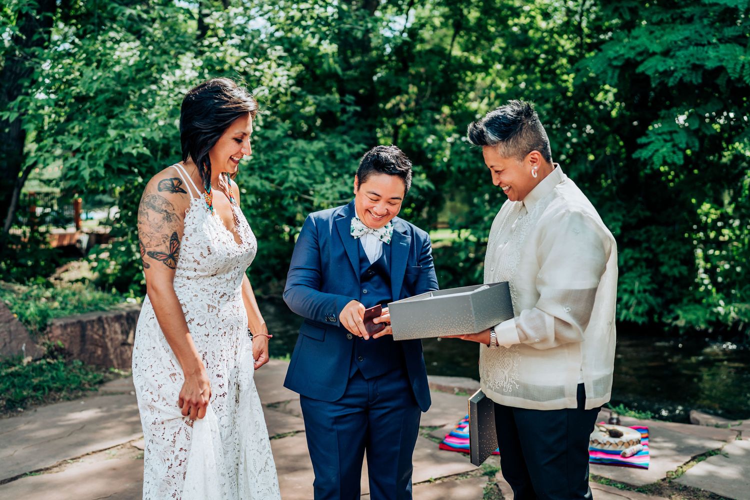 Three people gather during an outdoor ceremony, with one in a white lace dress and others in formal attire.