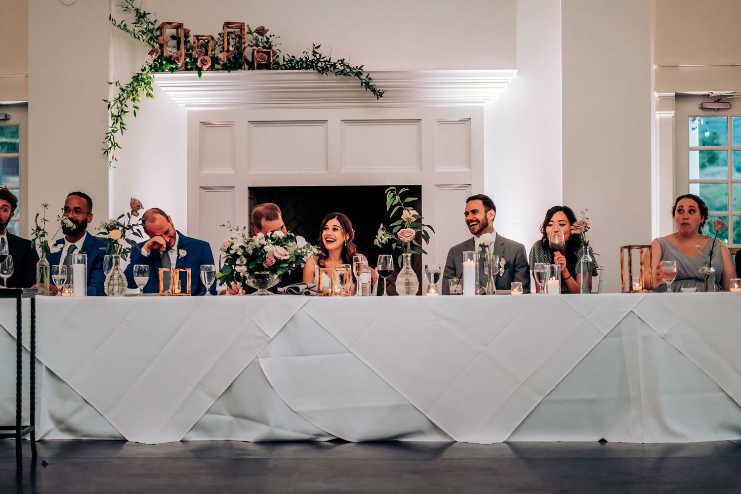 Wedding party seated at a long white head table decorated with greenery in a bright reception venue.