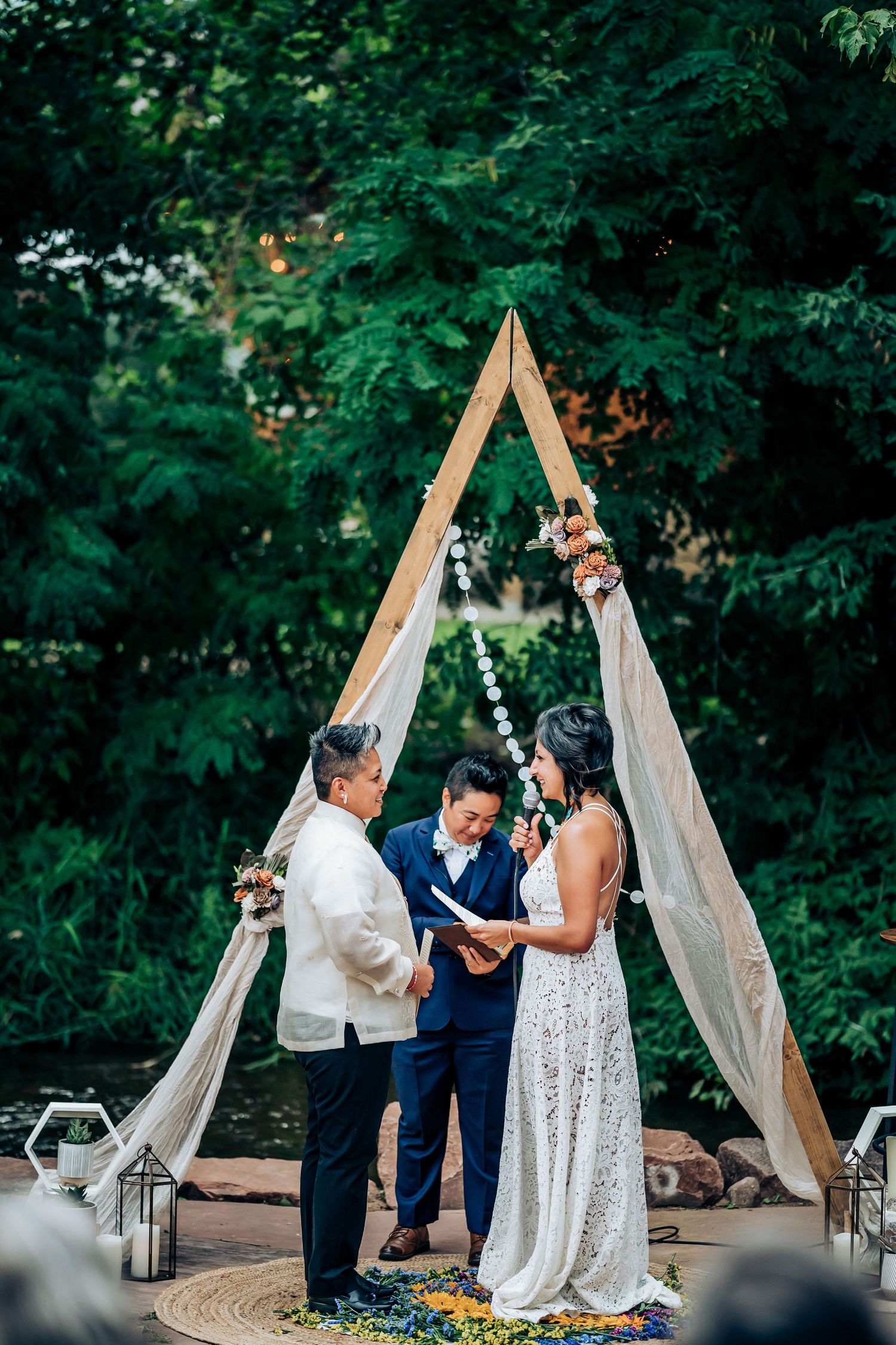 Wedding ceremony under a wooden triangle arch with draped white fabric against lush green foliage.