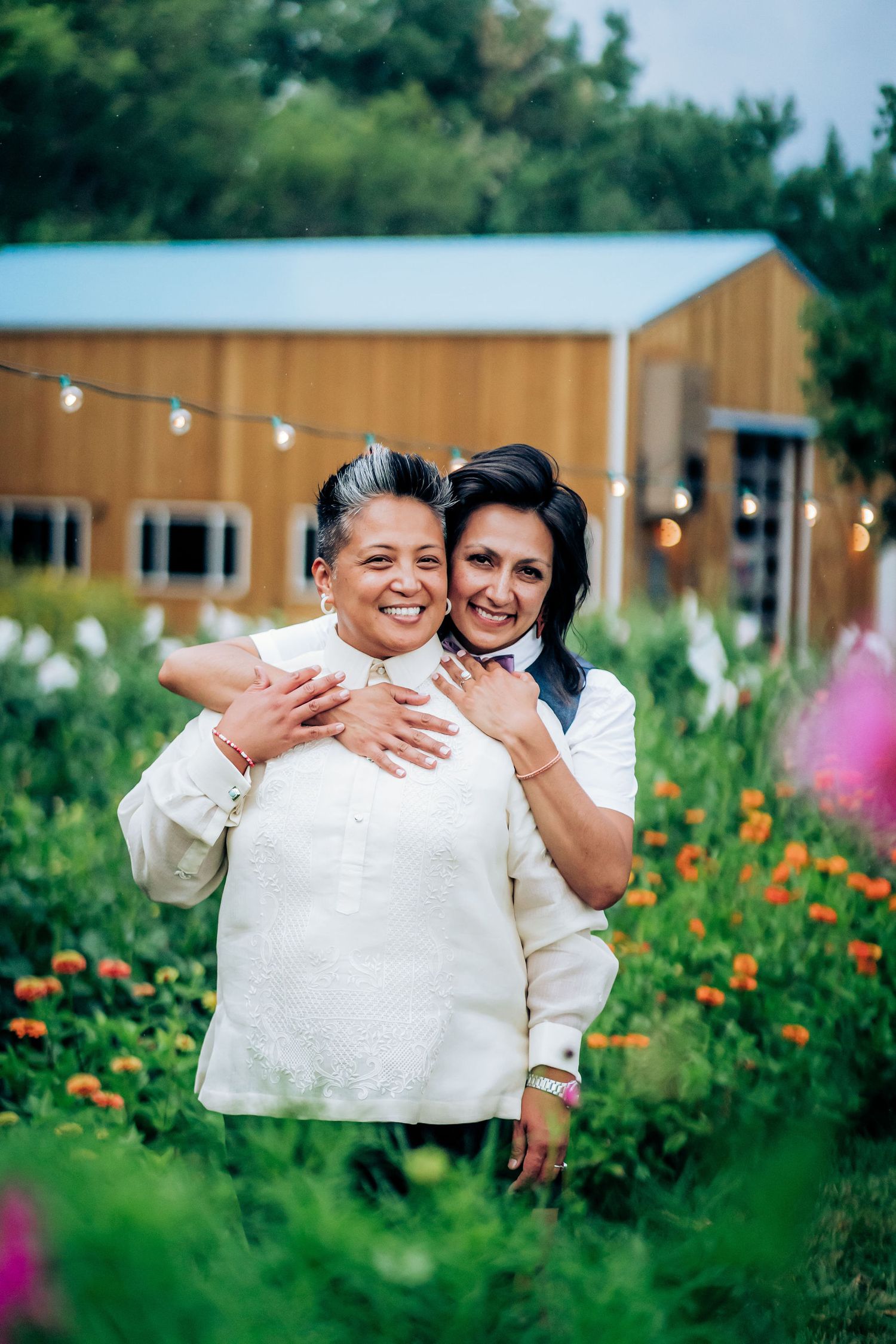 Two people embracing and laughing together in a garden with string lights and a barn in the background.