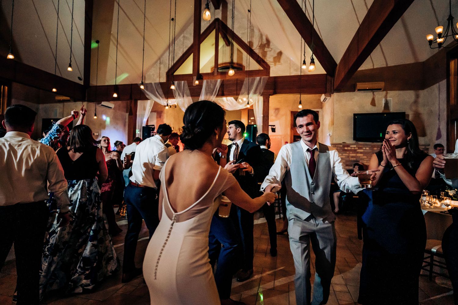 Wedding party dances in a rustic venue with exposed wooden beams and warm lighting.