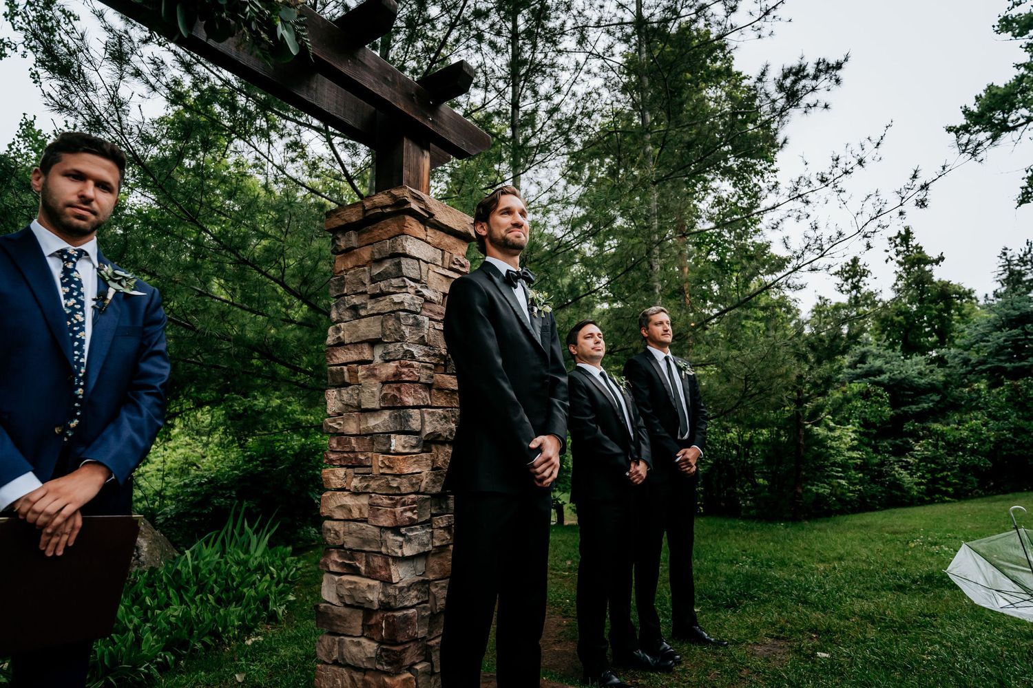 Groomsmen stand in formal attire near a stone pillar during an outdoor wedding ceremony.