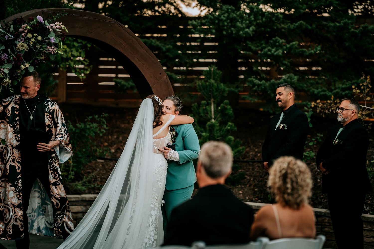 Bride in white gown and veil embraces partner in teal suit during outdoor wedding ceremony under circular arch.