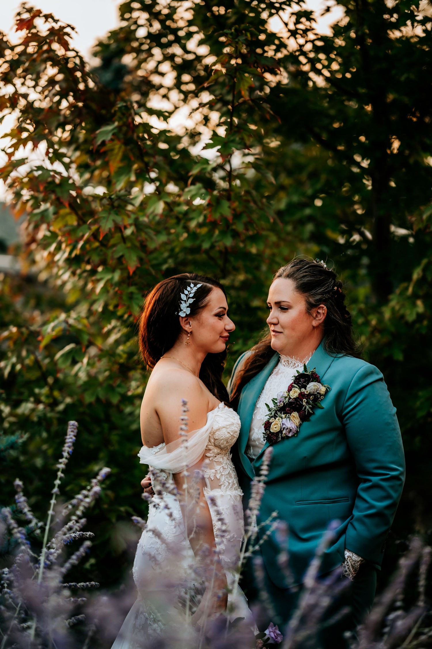 Two brides gazing at each other in a garden at sunset, one in a white gown and one in a teal suit.