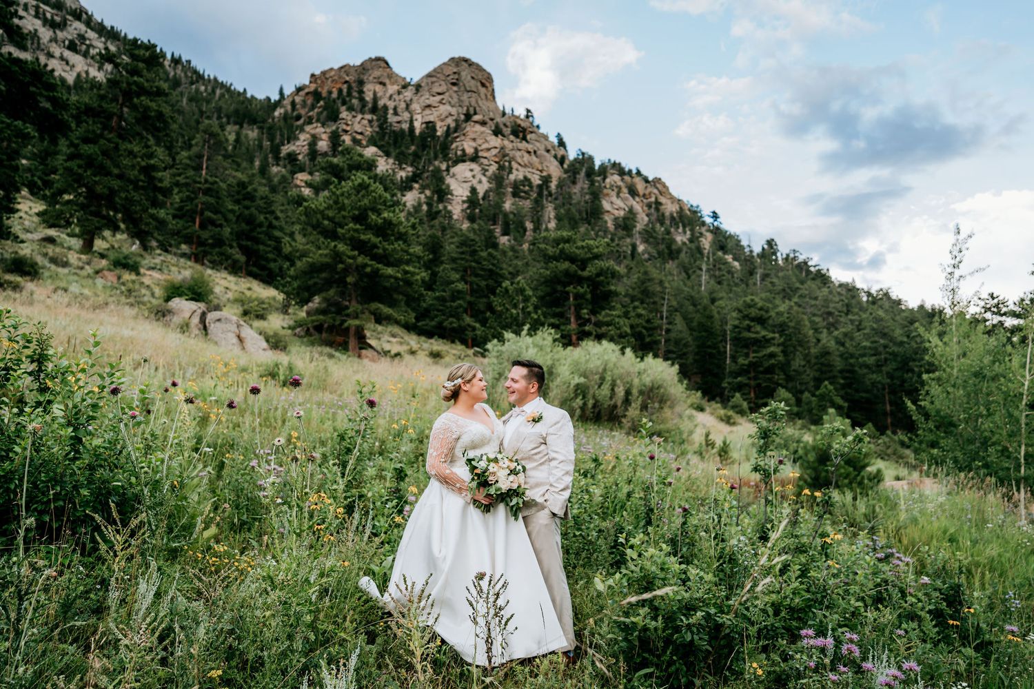 Scenic mountain wedding portrait in wildflower meadow with rocky cliffs in background.