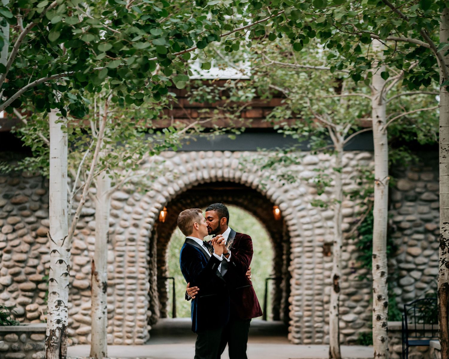 A romantic embrace under a stone archway surrounded by birch trees.