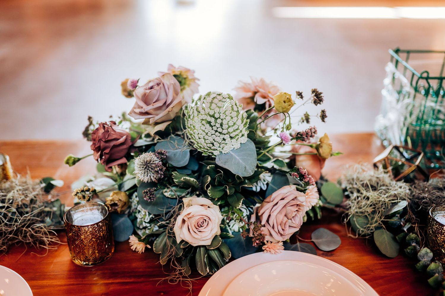 Romantic floral centerpiece with blush roses and greenery on wooden wedding reception table.