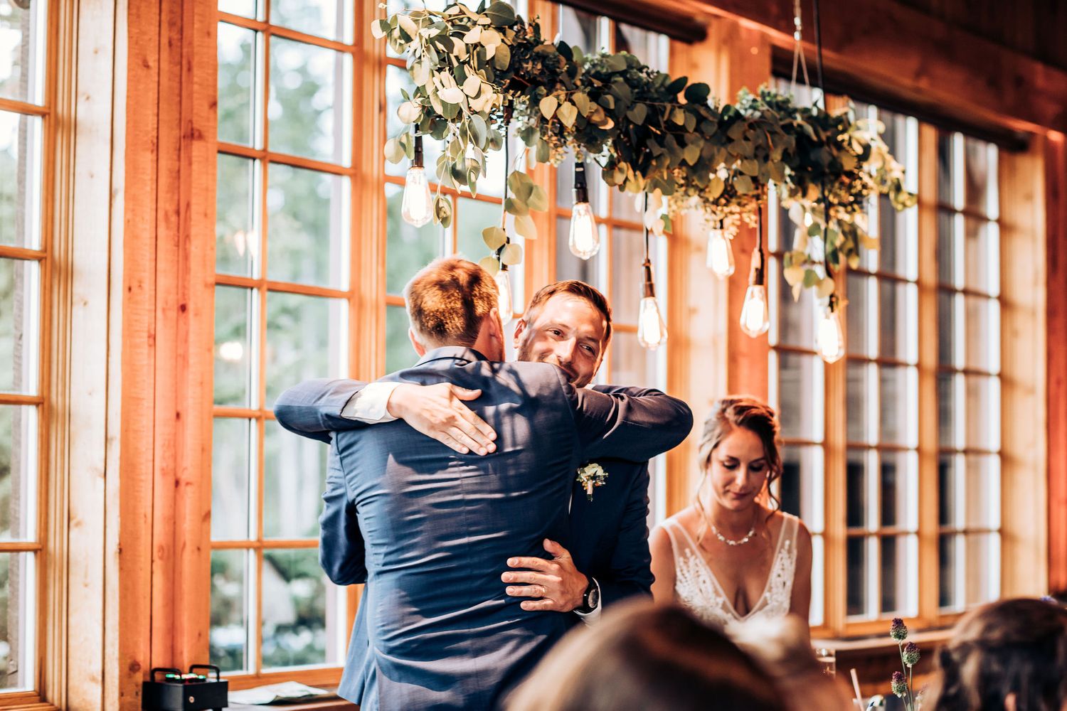 Emotional embrace during wedding reception in a sunlit rustic venue with greenery decor.