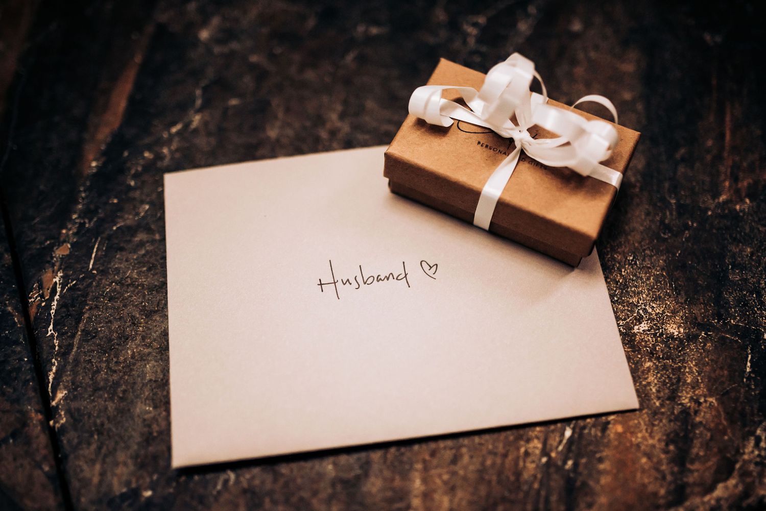 Wedding invitation and small gift box tied with white ribbon on rustic wooden surface.