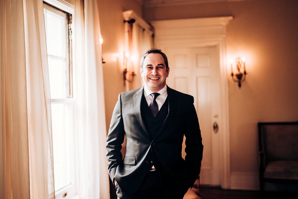 A groom in a formal dark suit poses by a window in a classical interior.