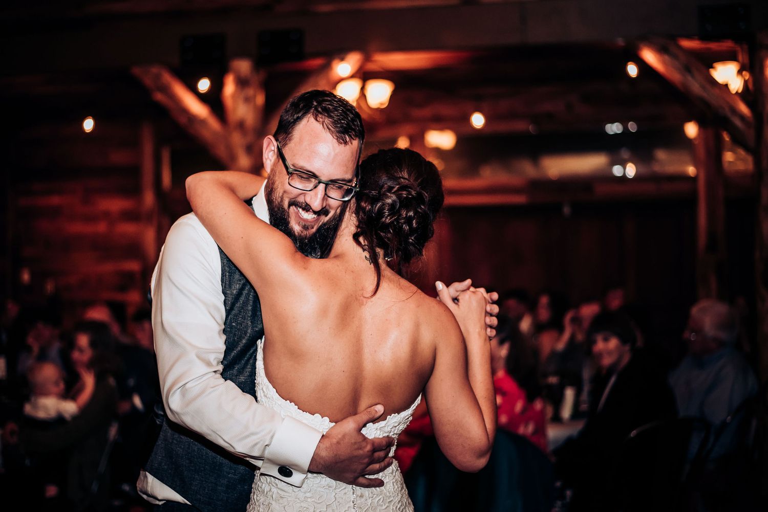 A tender slow dance moment between newlyweds in a warmly lit rustic reception venue.
