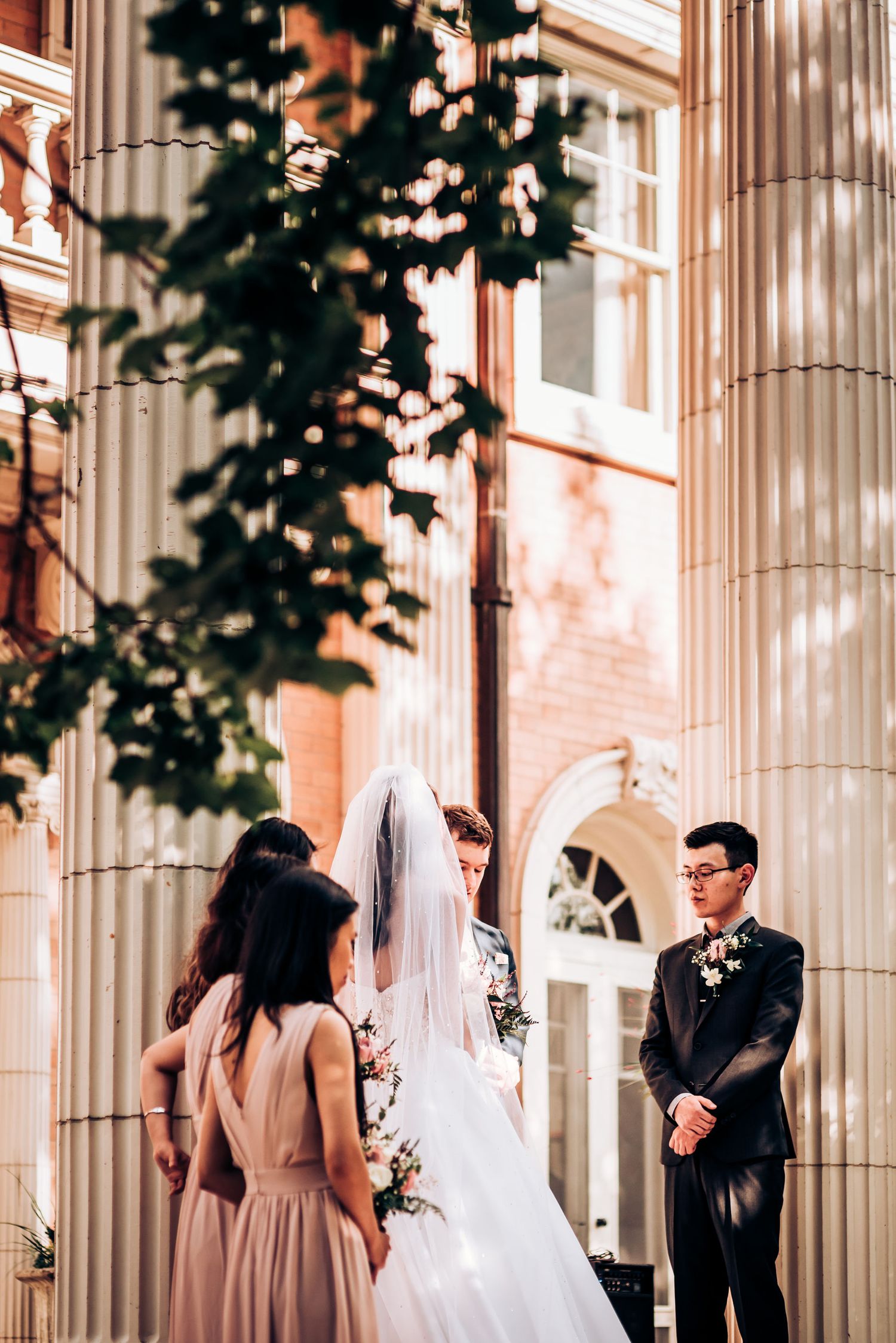 Wedding party gathers by classical stone columns during an outdoor ceremony.
