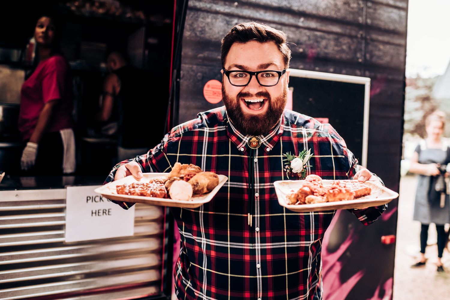 Smiling server in plaid shirt holds two plates of food at outdoor event.